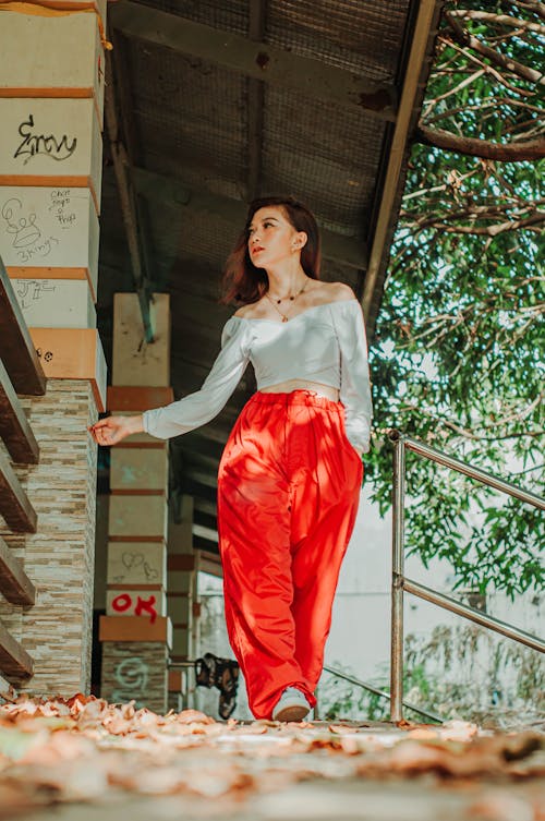 Full body of ethnic woman looking away against green trees while walking on porch with withered leaves