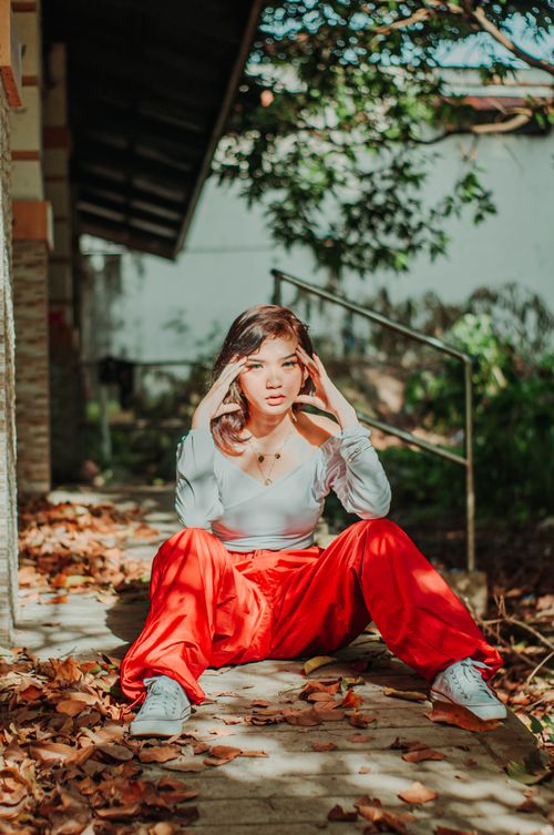 Free Trendy Asian female teenager in red pants touching temples while sitting among faded foliage and looking at camera Stock Photo