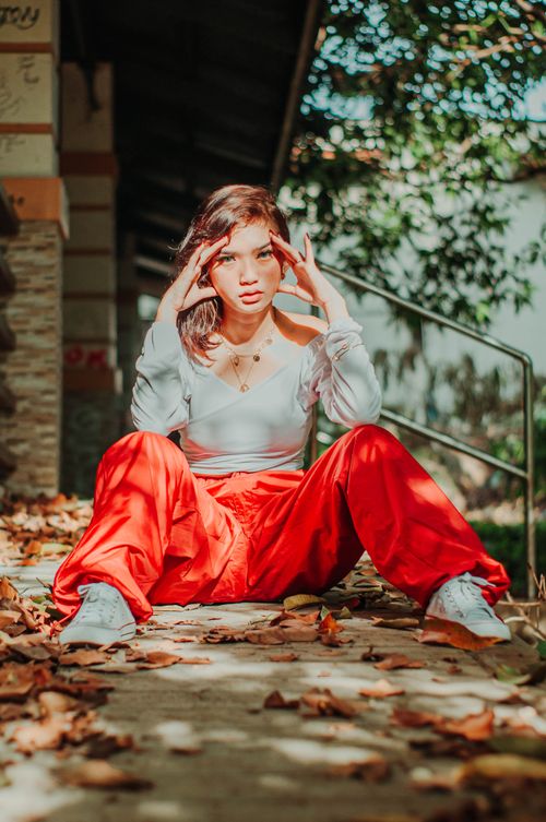 Free Trendy Asian female teenager in red pants touching temples while sitting on footpath and looking at camera in sunlight Stock Photo