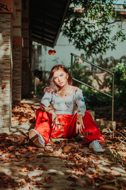 Free Full body of trendy millennial female in red pants looking at camera while sitting near house on ground with withered leaves Stock Photo