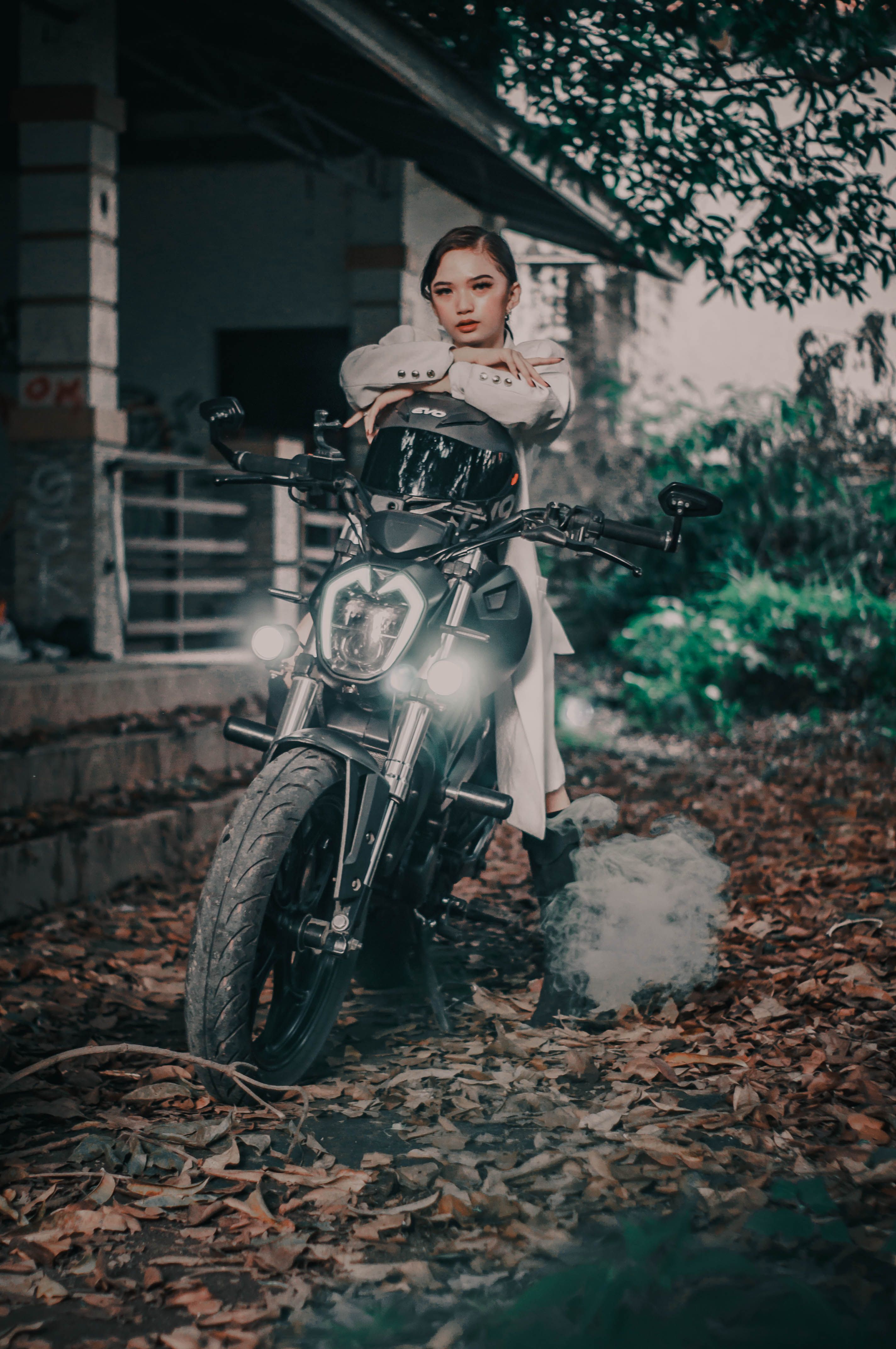 Full body of stylish brutal female sitting on motorbike with headlight near weathered house with fence on ground with weathered leaves in autumn day