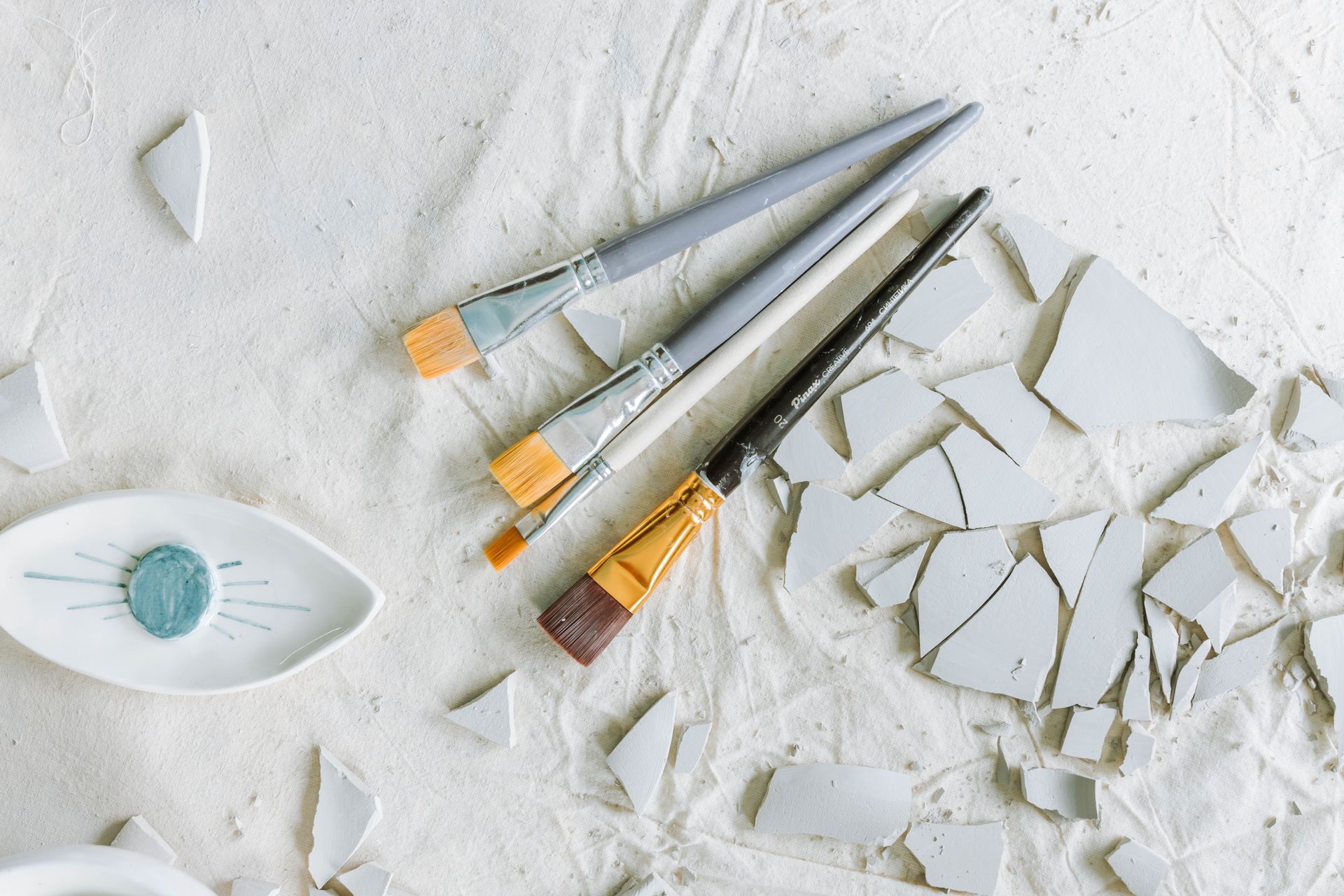 Close-up of paint brushes with broken ceramic shards on a light canvas.