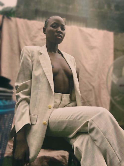 Self esteem young fit African American woman with short hair wearing trendy suit on naked torso sitting on chair on yard of aged village house