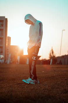 A young man in sportswear and hoodie standing on a field during sunset, exuding a sporty vibe.