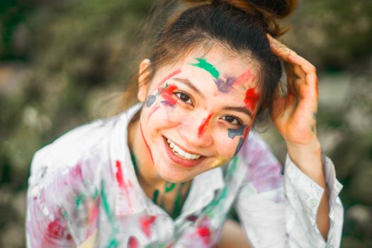 Cheerful young ethnic female with black hair tied up and multi colored paint flecks on face looking at camera with bright smile in park on blurred background