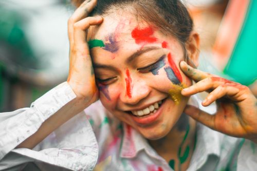 Free Cheerful young ethnic female with dye on face touching forehead and cheeks with eyes closed on blurred background Stock Photo