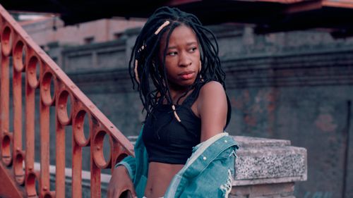 Free Young woman with dreadlocks stands by metal railing outdoors, exuding style in denim and crop top. Stock Photo