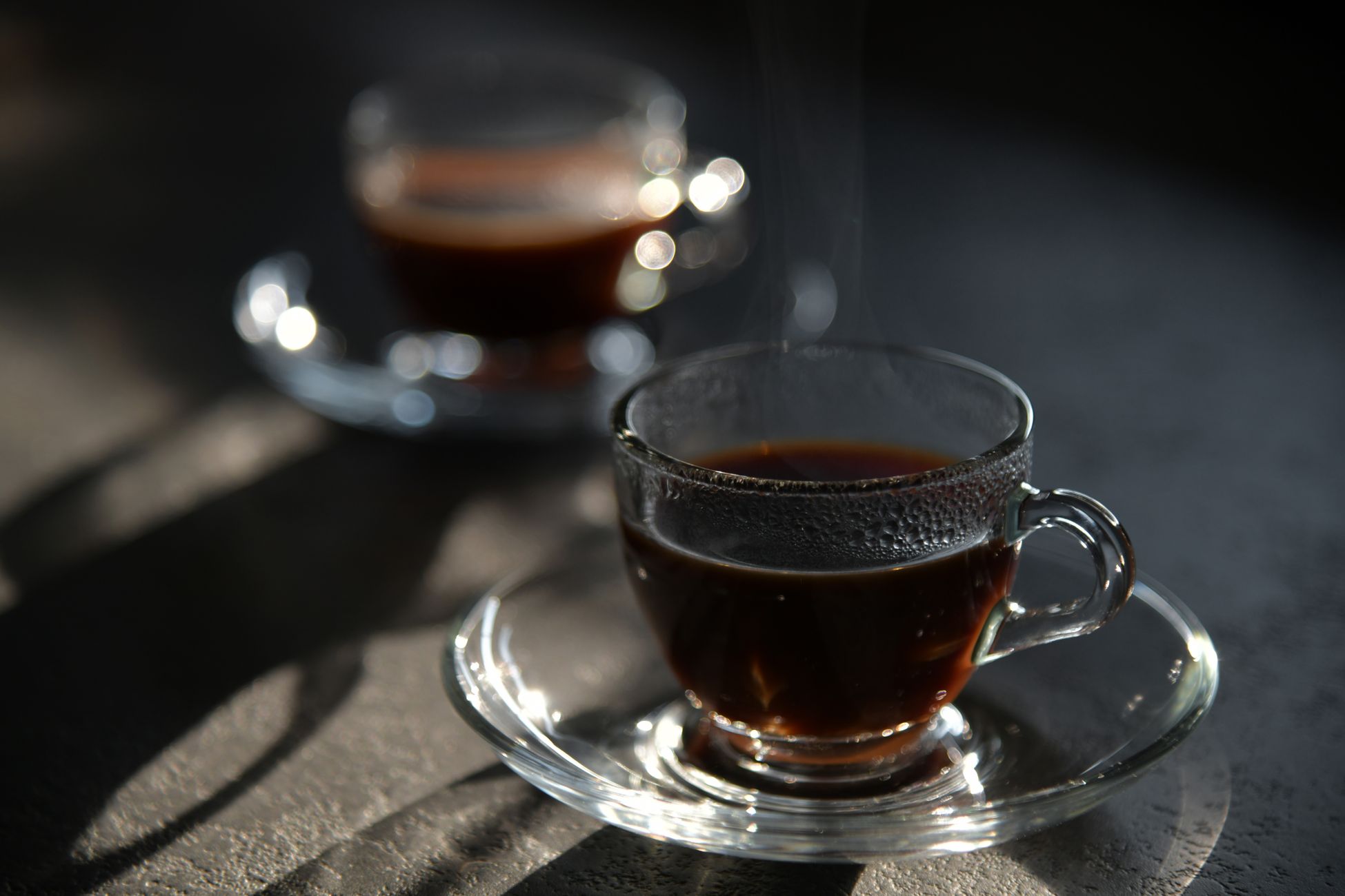 Close-up of steaming black coffee in elegant glass cup with saucer, captured in soft light.