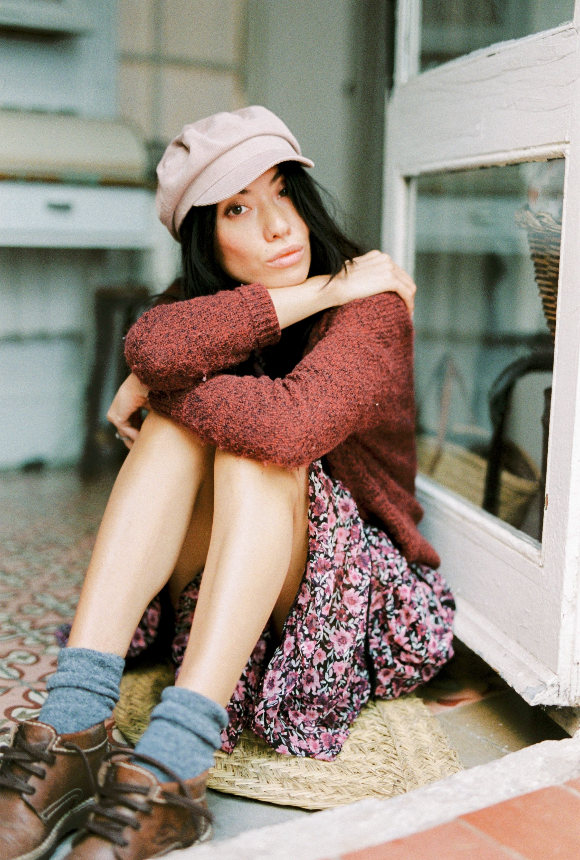 Young woman sitting indoors, wearing a knit sweater and floral skirt, exuding a relaxed vibe.