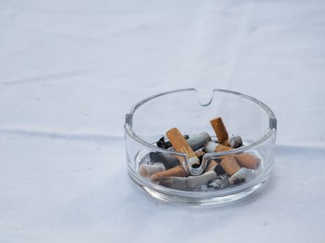 A clear glass ashtray filled with cigarette butts and ash placed on a white surface.