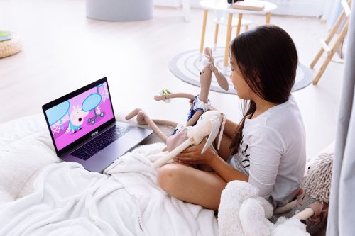Free Young girl holding plush toys while watching a cartoon on a laptop, sitting on a cozy bed. Stock Photo