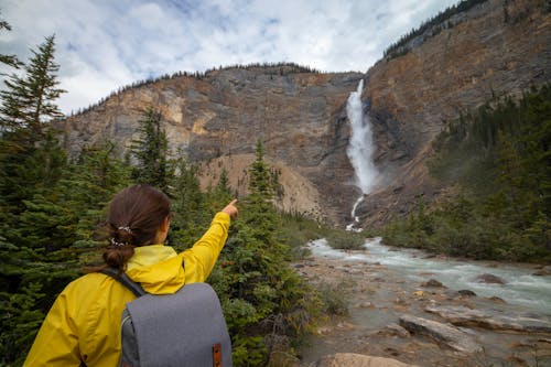 Woman in Yellow Raincoat Pointing a Finger to Waterfall in Mountains