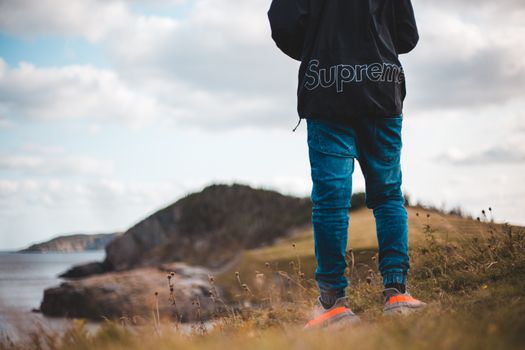 Back view of crop anonymous male tourist on grassy mount near ocean under cloudy sky