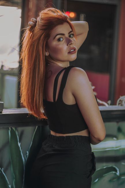 Side view of self assured young lady in casual clothes touching long red hair and looking at camera while relaxing in bar