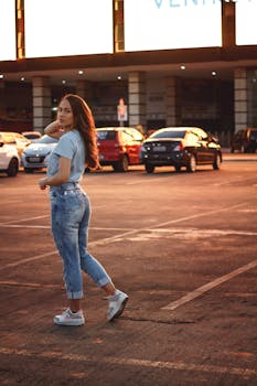 Side view of confident ethnic female teenager with long brown hair in trendy outfit walking along parking lot at sunset and looking at camera
