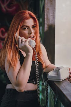 Smiling young woman with red hair chatting on a vintage rotary phone indoors.
