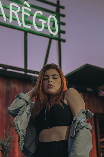 Low angle of sensual young lady in trendy crop to and denim jacket touching long ginger hair and looking at camera while relaxing on city street against cloudless purple sky