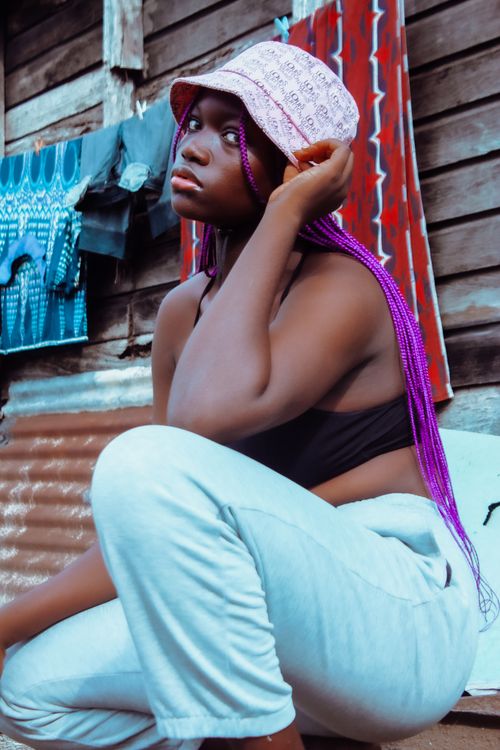 Free A fashionable woman with purple dreadlocks and a bucket hat posing outside against a rustic backdrop. Stock Photo