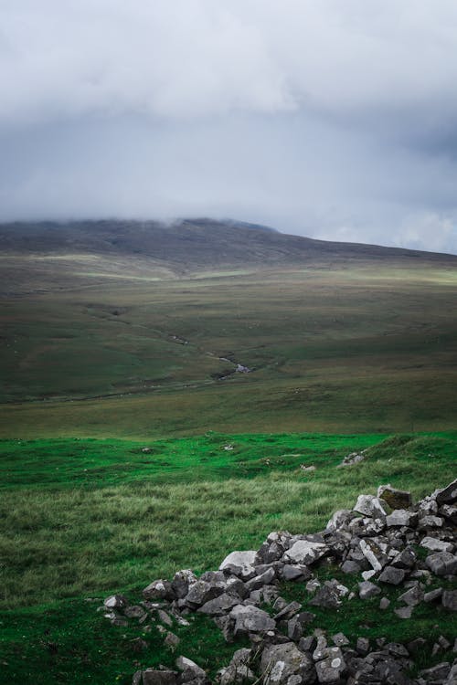 Free Picturesque scenery of green grassy hill and stones on surface under cloudy sky Stock Photo