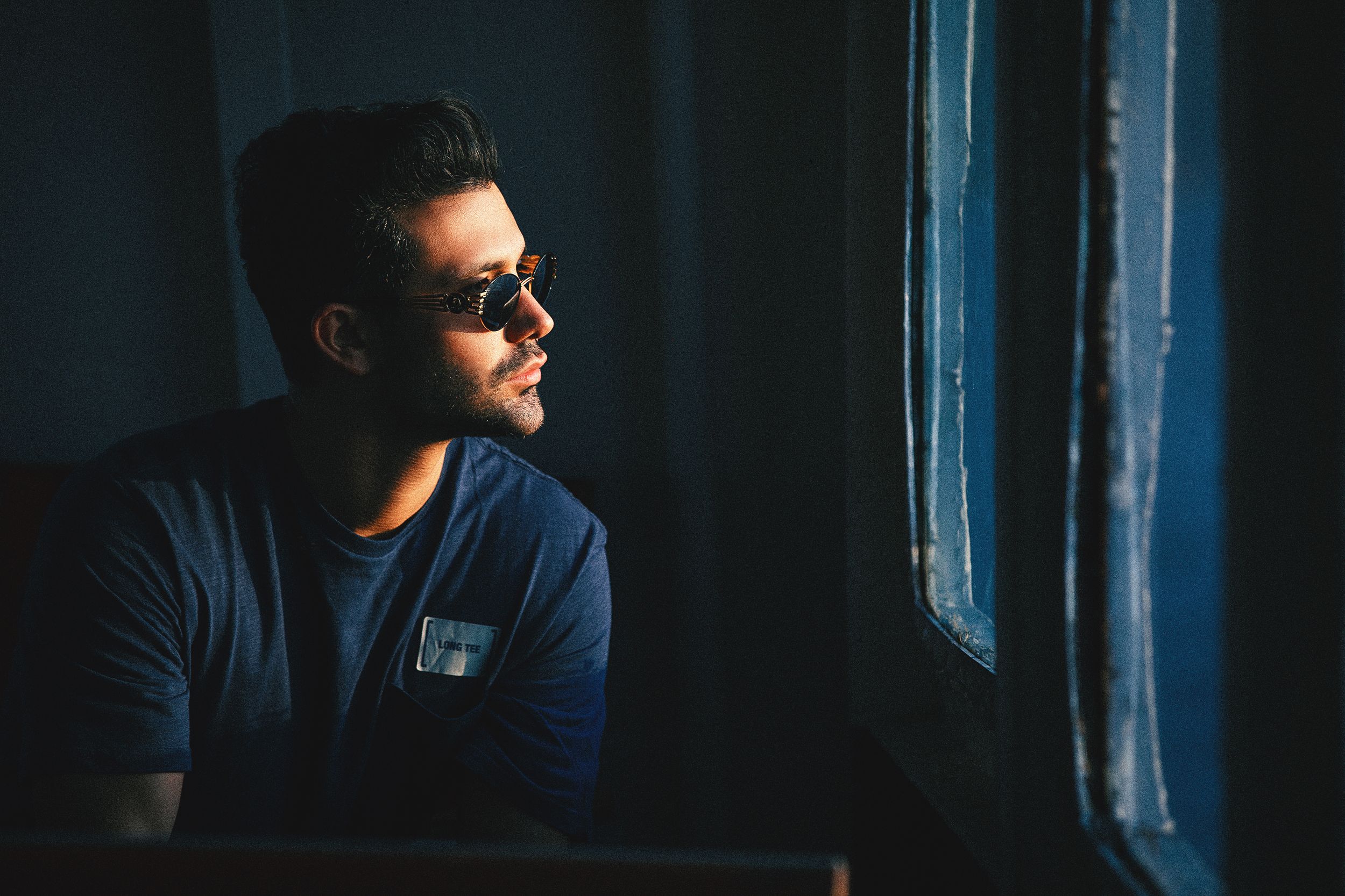 Man in sunglasses and blue shirt gazing thoughtfully out a window in low light.