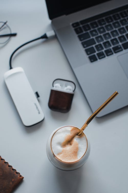 From above glass of coffee with foam and zero waste straw placed on table with laptop and USB hub and earphones