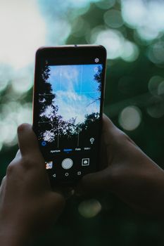 Capture of a smartphone taking a photo of the sky through trees outdoors.