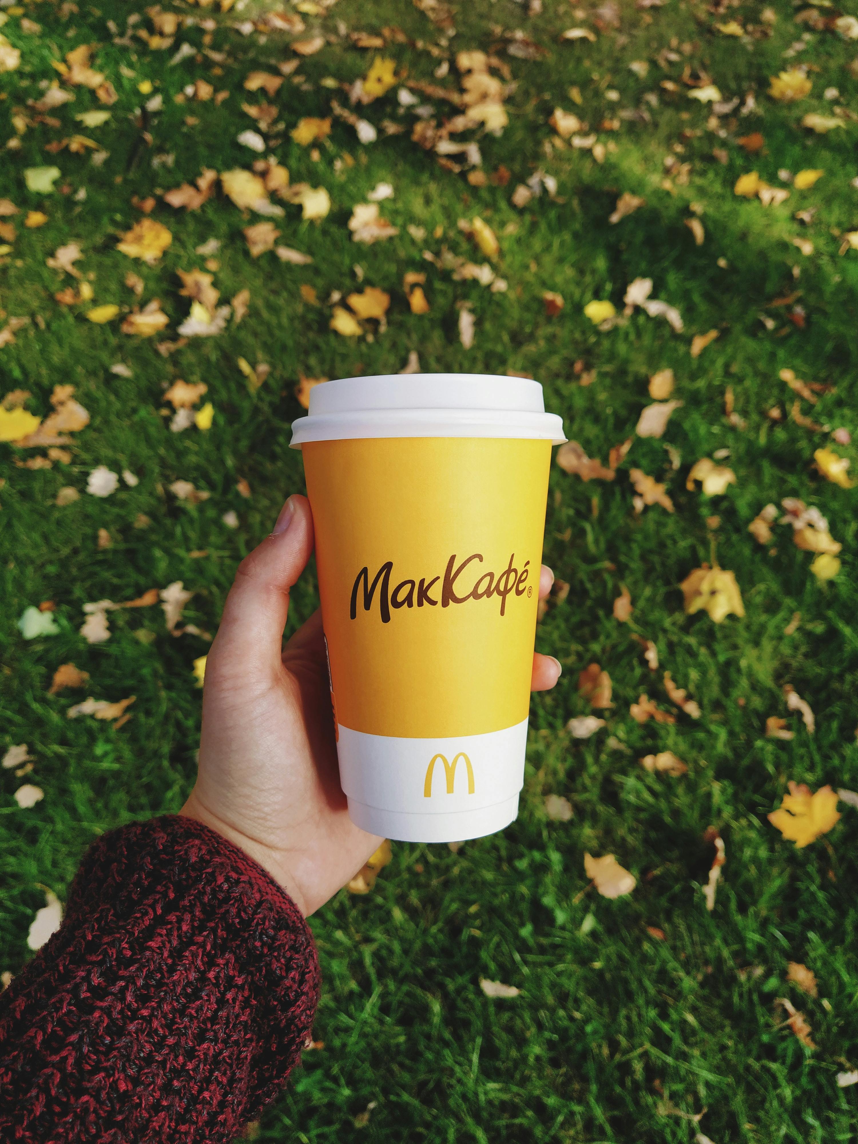 Hand holding McCafé cup in autumn park with fallen leaves on grass.