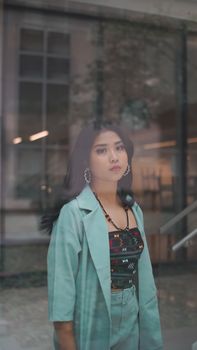 An Asian woman in fashionable attire stands by a glass window, showcasing a modern style.