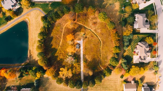 Top view of countryside area with trees near artificial pond and building roofs in autumn