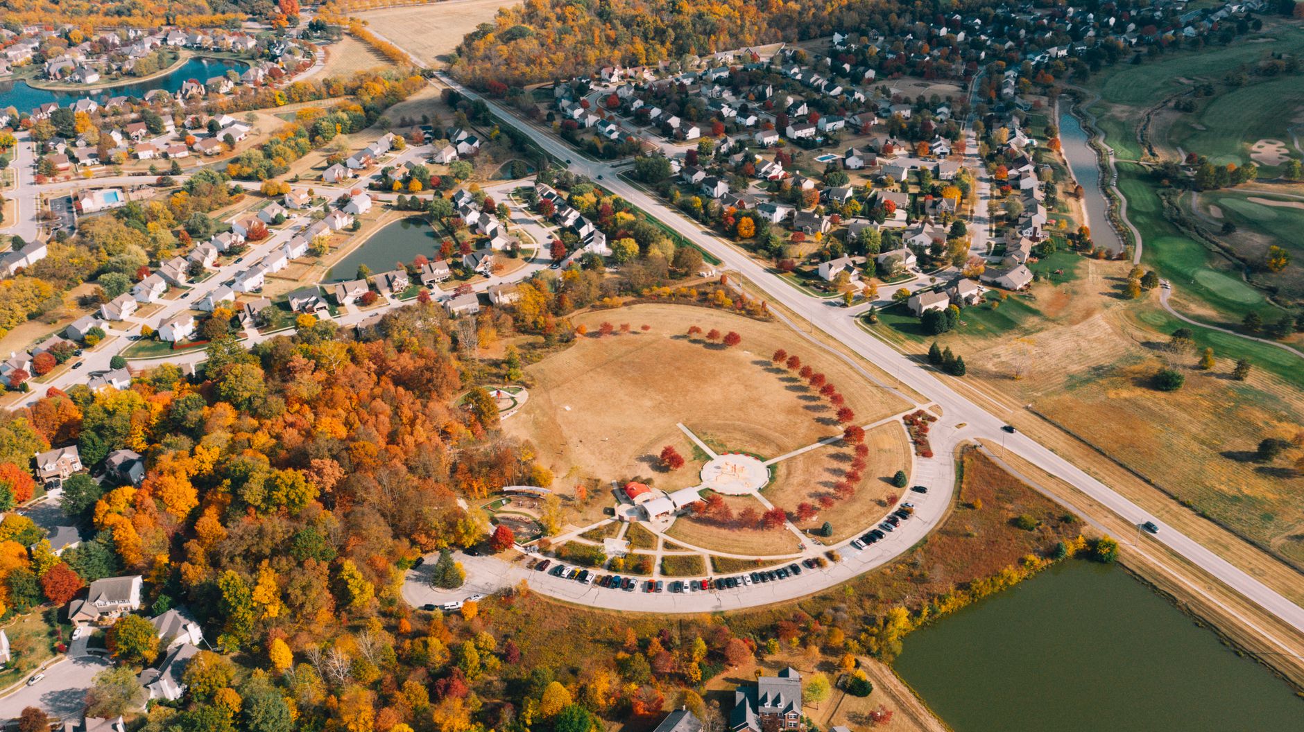 Drone view of bright lawn with colorful trees near residential building roofs and water channel with road in fall