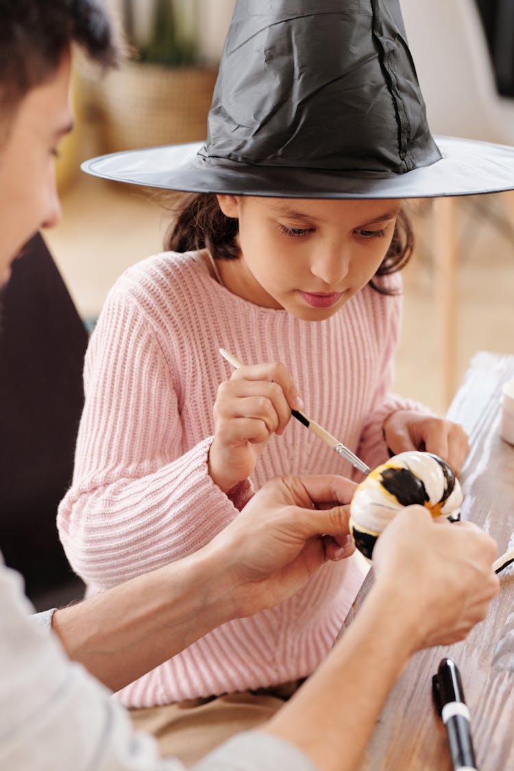 A Girl With A Witch Hat Painting A Pumpkin With Black And White