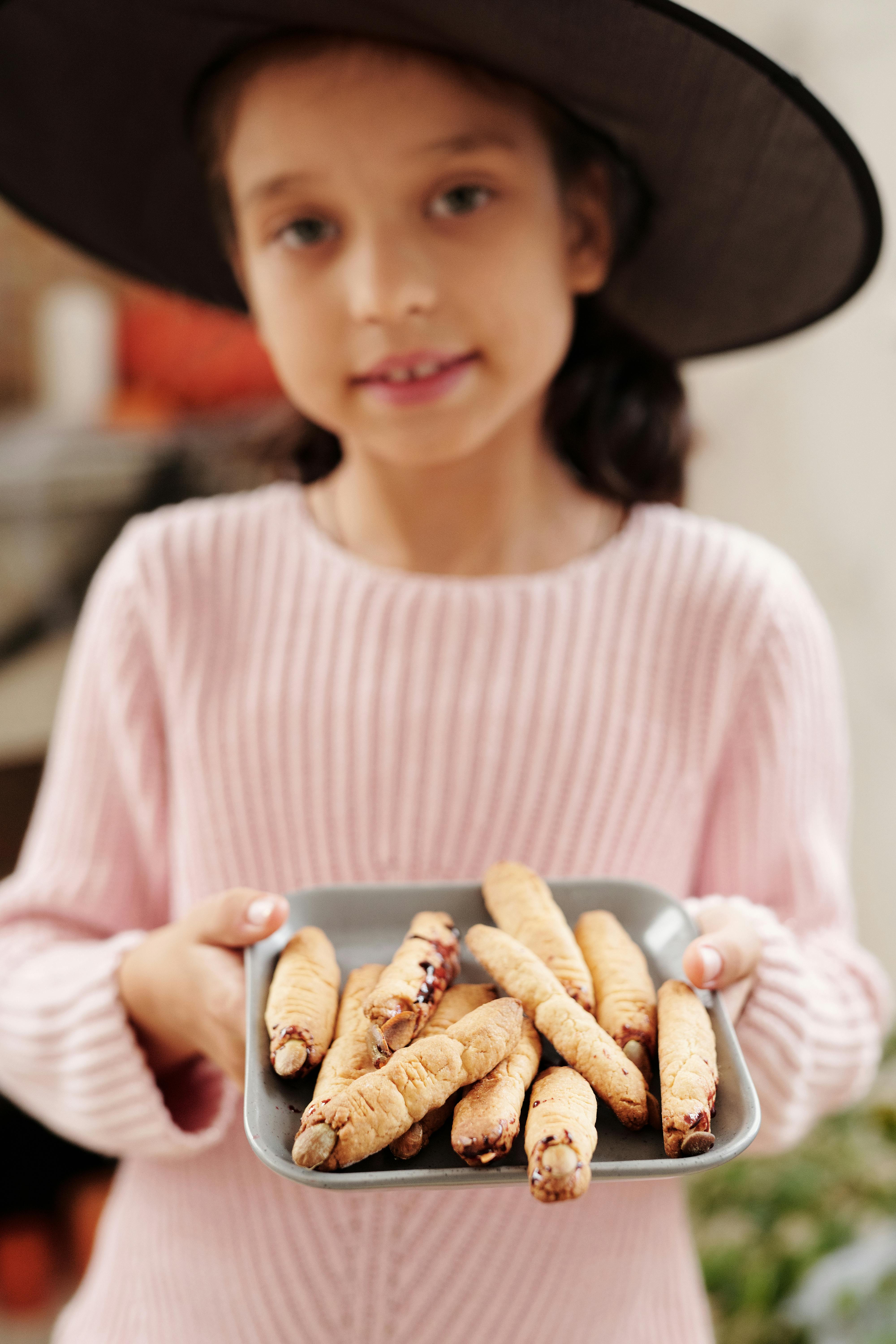 A young girl in a witch hat holding a tray of Halloween-themed witch finger cookies indoors.