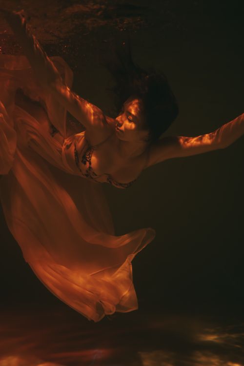 Free Artistic underwater photograph of a woman in a flowing dress, captured in warm, orange light for a dreamlike effect. Stock Photo