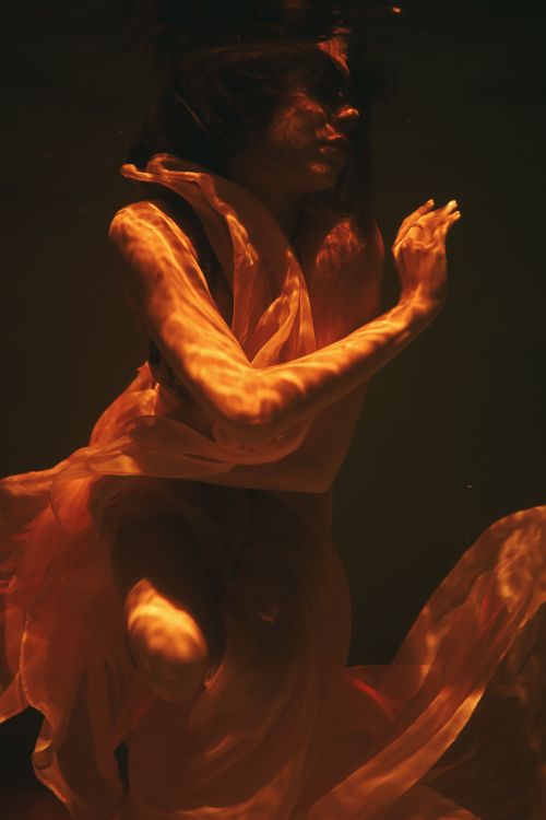 Free A graceful woman submerged underwater with sheer fabric, illuminated by orange light. Stock Photo