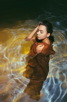 一名身穿棕色连衣裙的女子，正闭着眼睛，在波光粼粼的水中平静地摆着姿势