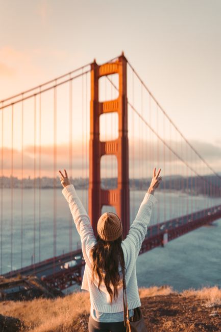Woman celebrating with peace sign gestures at Golden Gate Bridge at sunset.