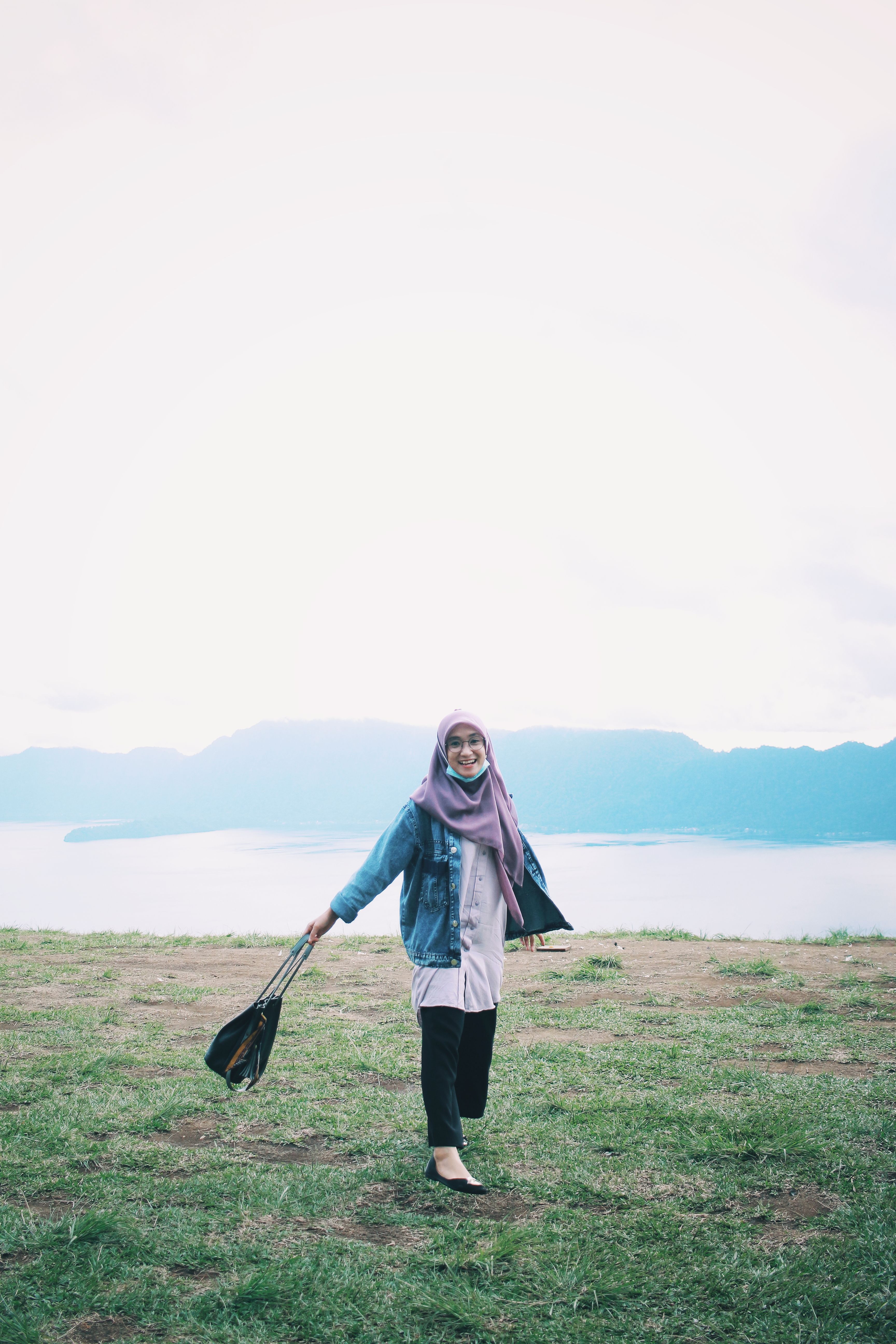 A joyful woman in hijab smiles standing on grassy lakeside with mountains in the background.