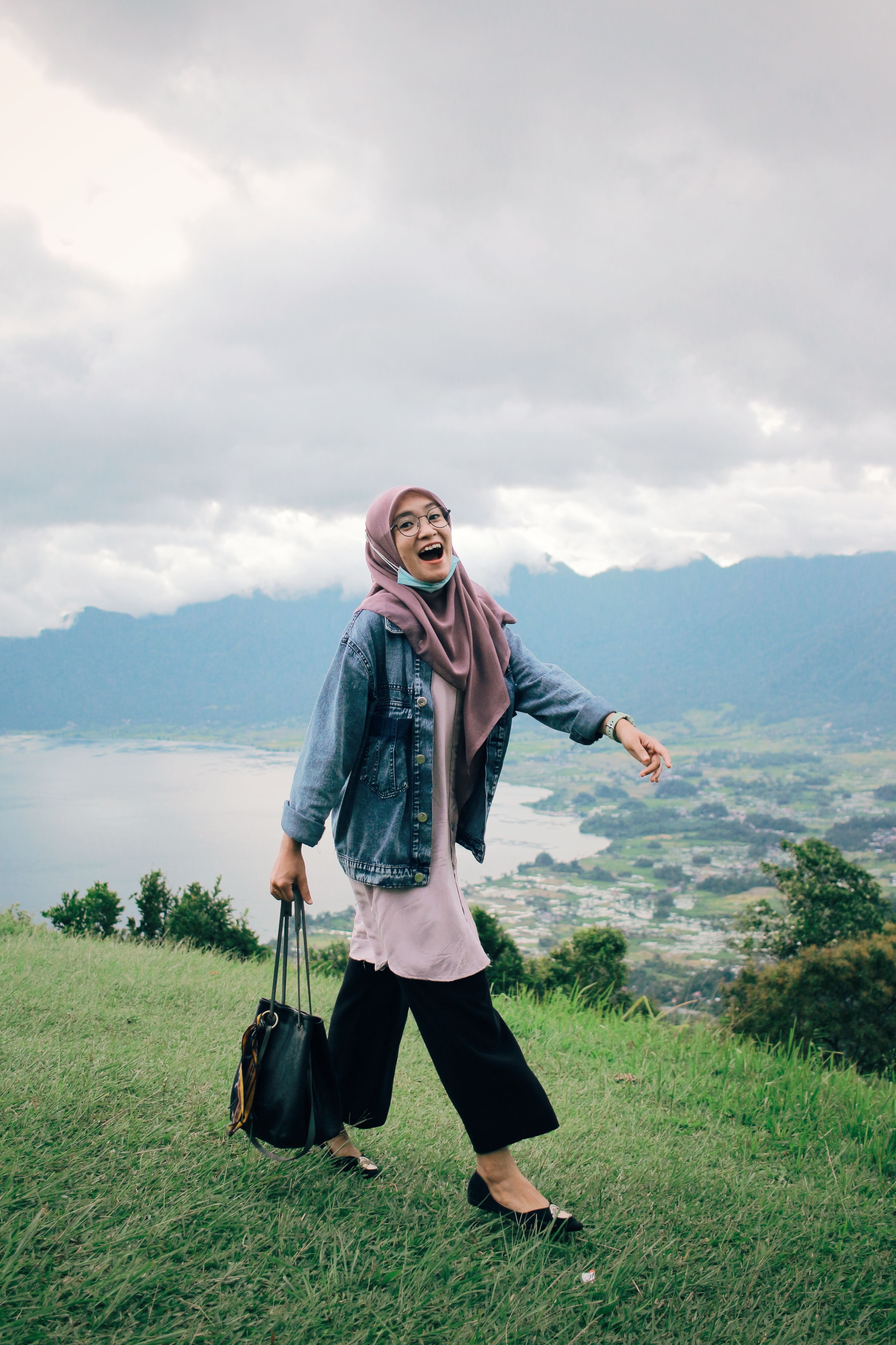 Woman in a denim jacket and hijab smiles while walking on a hill with a mountain backdrop.