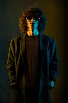 一位卷发、戴着太阳镜和外套的时尚男子肖像，在戏剧性的灯光下，散发着神秘与风格