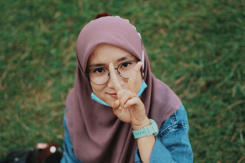 Free Asian woman wearing hijab and glasses, posing with peace sign on grass. Stock Photo
