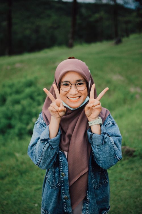 Free Cheerful woman in hijab, denim jacket, and glasses posing gleefully with peace signs outdoors. Stock Photo