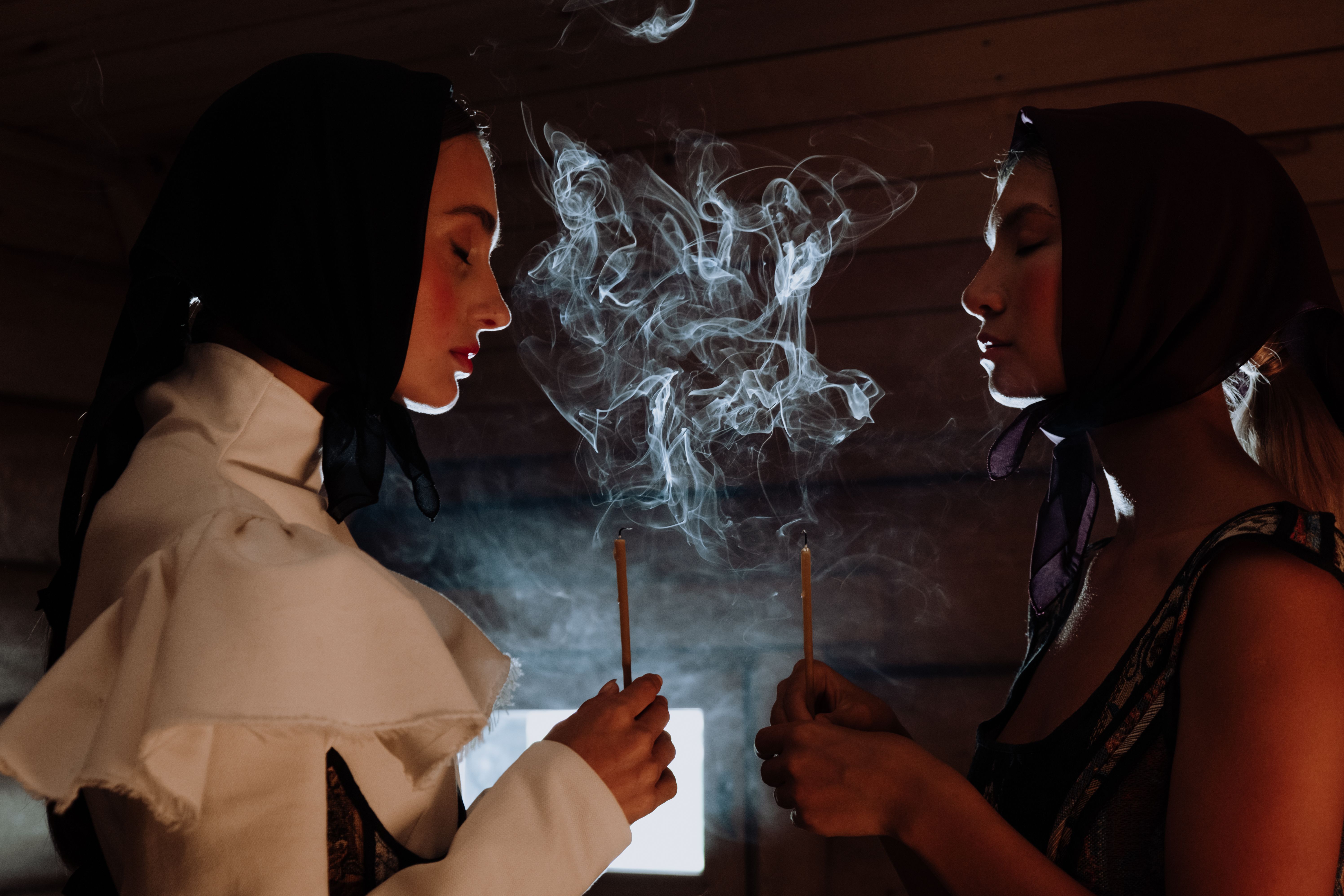 Two women in headscarves hold incense sticks with ethereal smoke indoors, creating a mystical atmosphere.