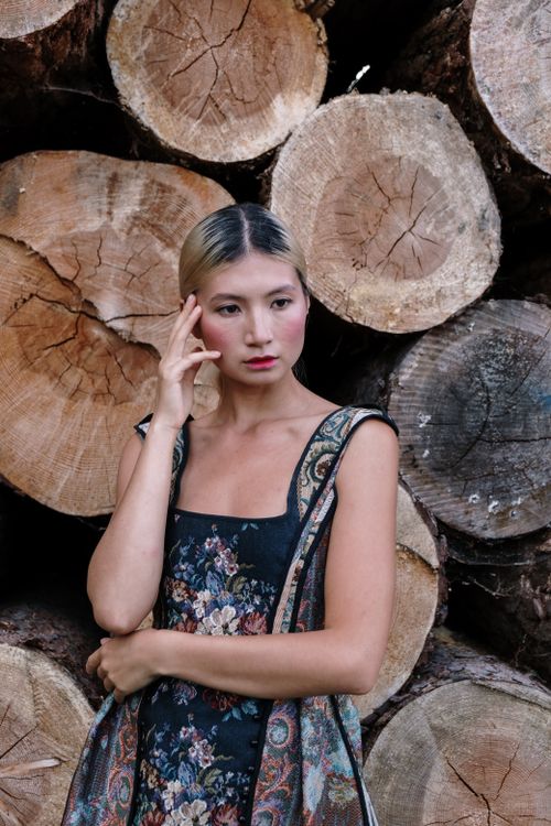 Free Stylish Asian woman poses in a floral dress against a rustic wooden log backdrop. Stock Photo