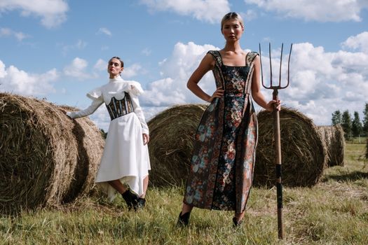 Two women in stylish dresses posing with haystacks in a rural setting.
