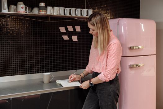 Caucasian woman standing beside pink refrigerator taking notes in a modern kitchen.