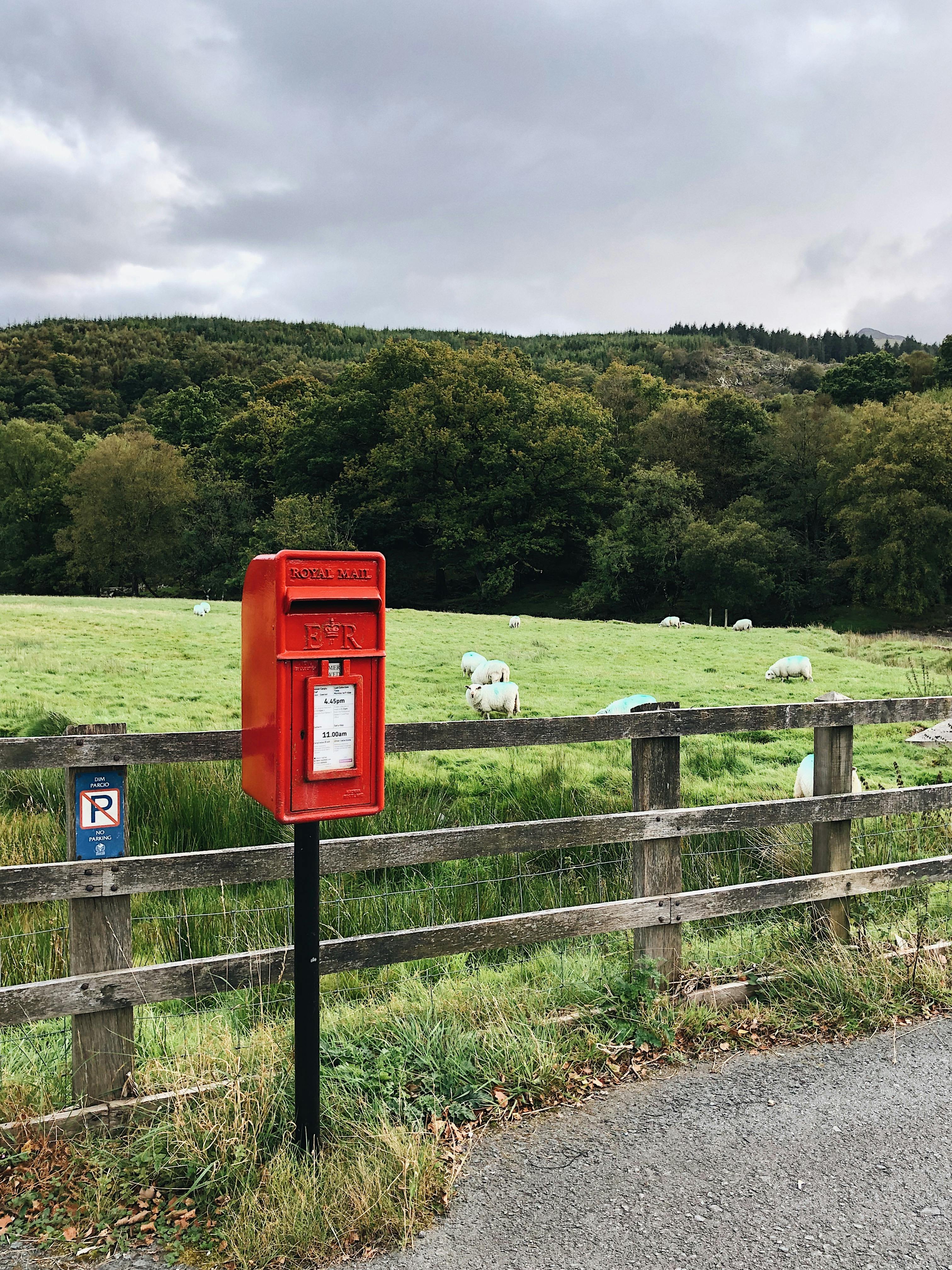 A vibrant red Royal Mail postbox set against a rural Welsh countryside landscape.