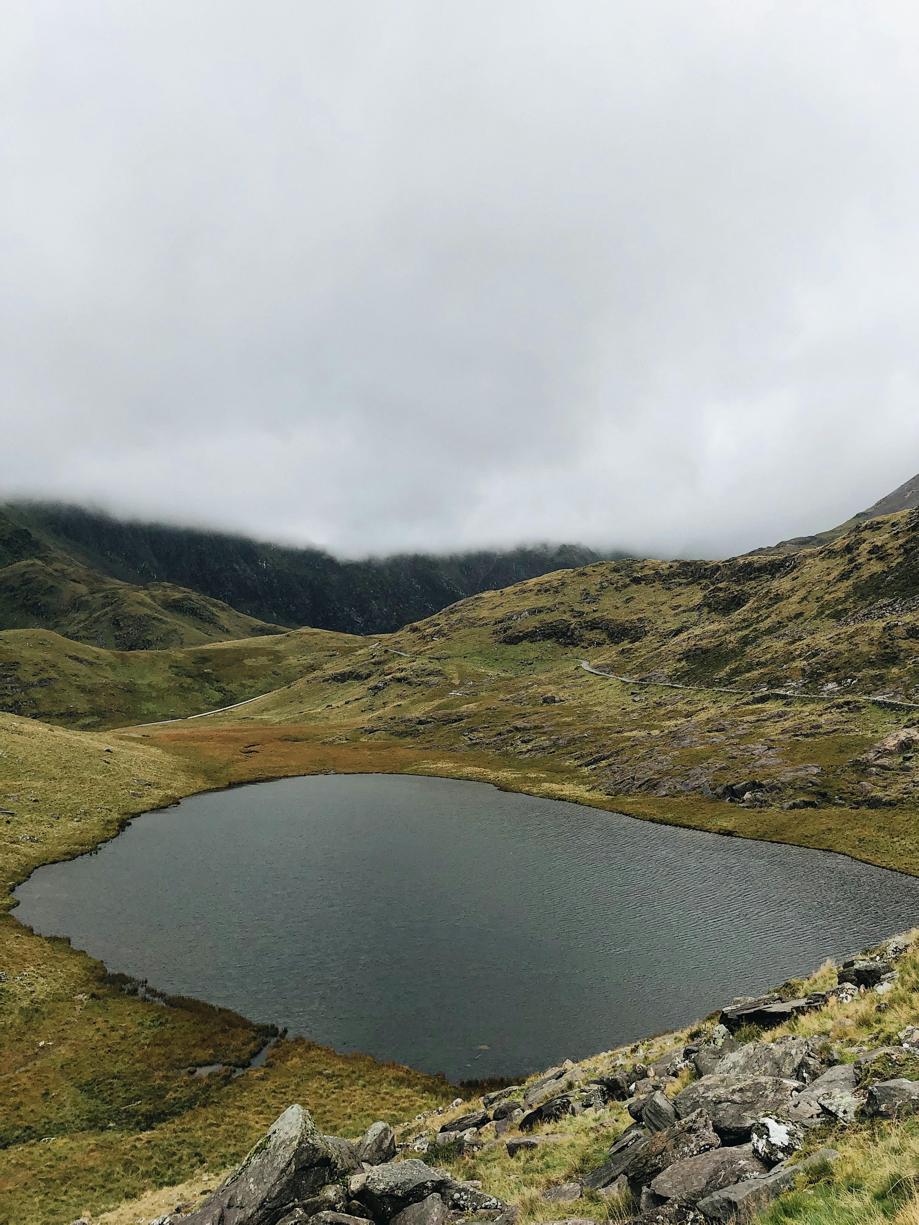 A serene landscape featuring a tranquil lake amidst rolling hills in Gwynedd, Wales, under a cloudy sky.