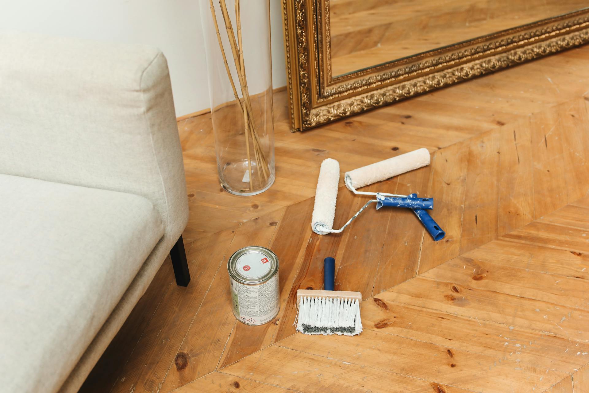 Paint supplies laid out on a wooden floor beside a couch and a large framed mirror, ready for home renovation.