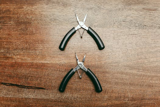 Close-up of two needle-nose pliers on a wooden tabletop, showcasing stainless steel construction and black handles.