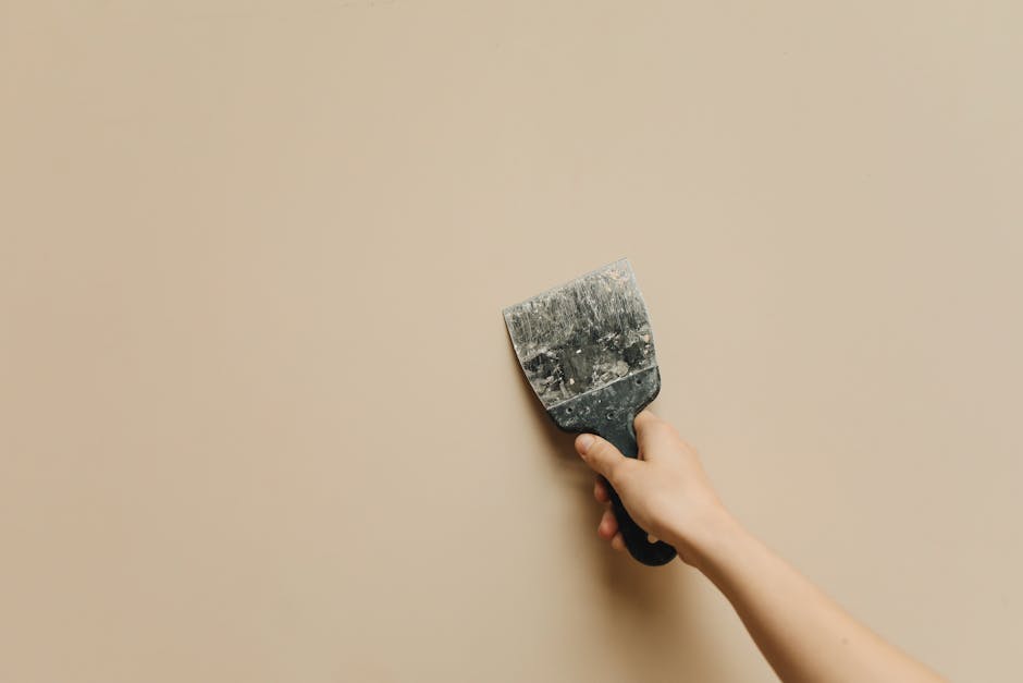 No Repairs Needed - cash home buyers in houston Hand holding a putty knife against a beige wall, illustrating the concept of home repairs and renovations relevant to selling properties as-is for cash.